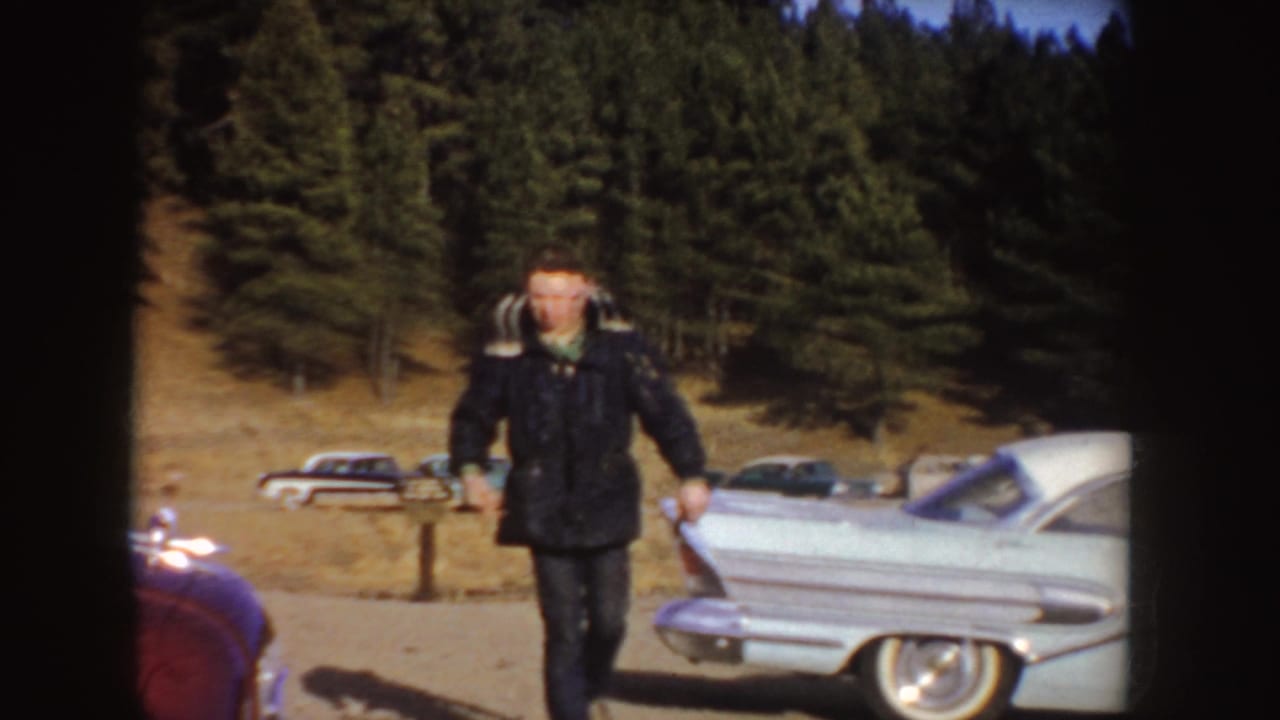 A man on roller skates approaches near parked midcentury cars with a wooded Idaho hillside in the background.