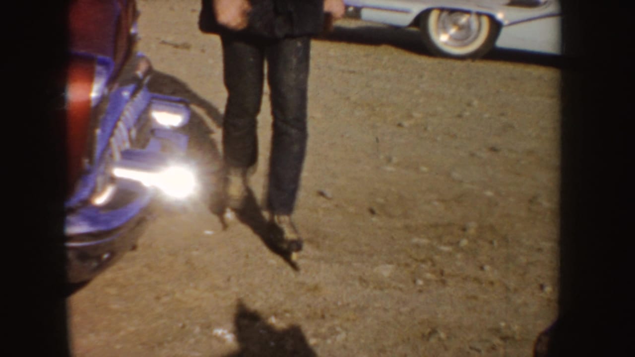The man’s roller skates and legs are visible beside parked cars on a sunlit dirt roadside in the 1955 Idaho clip.