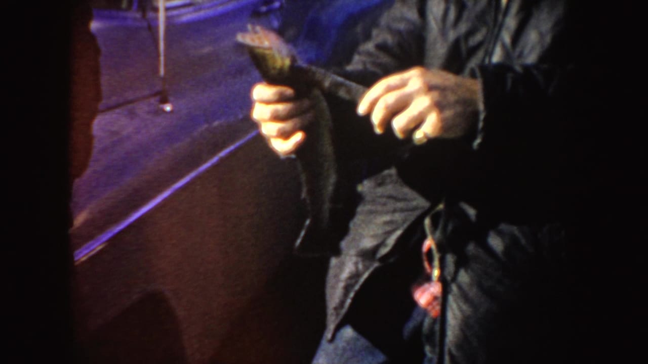 The man continues holding the fish near a vintage vehicle, with the frame cropped around his jacket, hands, and the catch.