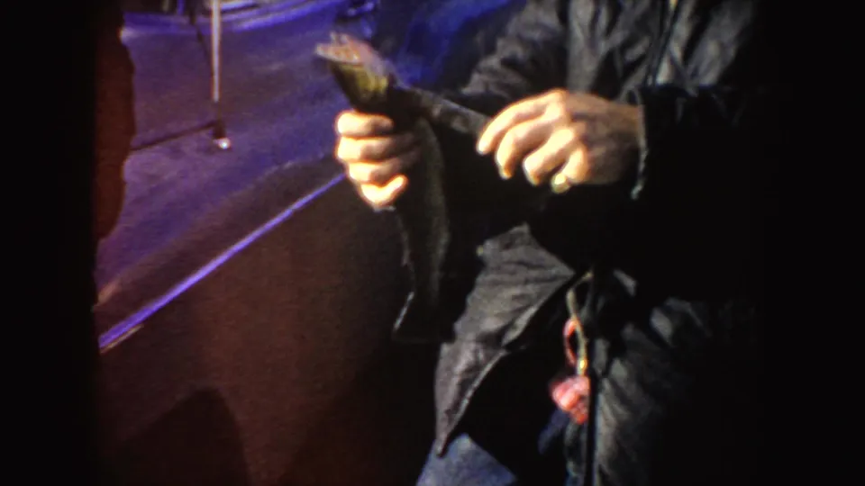 The man continues holding the fish near a vintage vehicle, with the frame cropped around his jacket, hands, and the catch.