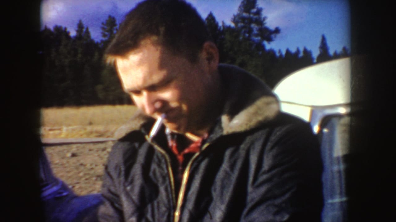 The man is framed in daylight with a cigarette, forest background, open ground, and a pale camper or vehicle surface at the right edge.