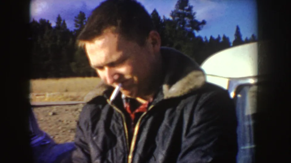 The man is framed in daylight with a cigarette, forest background, open ground, and a pale camper or vehicle surface at the right edge.