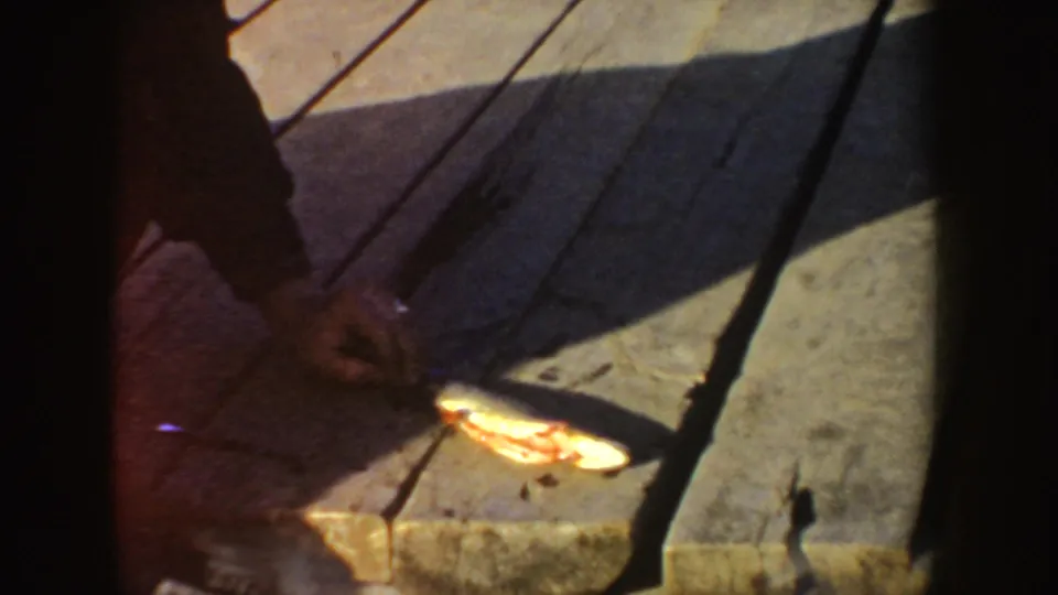 1955 Idaho home-movie frame showing a fish on wooden dock planks while hands work nearby in shadow.
