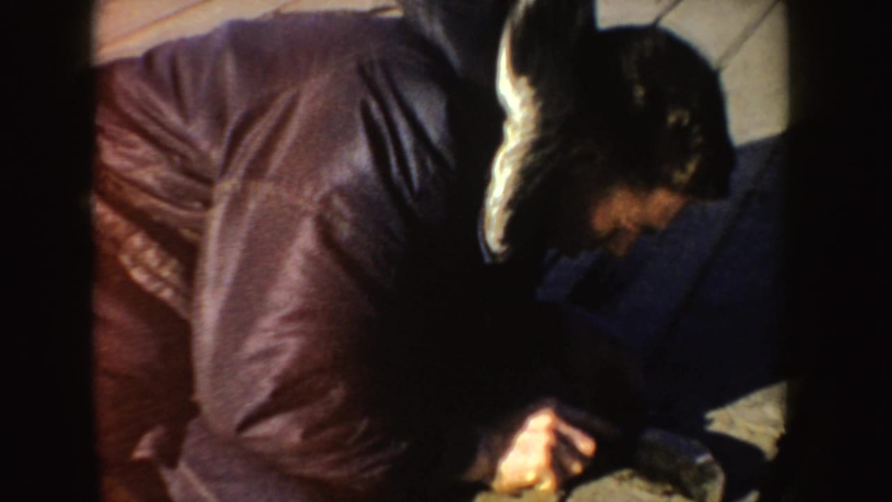 Man in a dark winter coat leans over a wooden dock surface in 1955 Idaho home-movie footage.