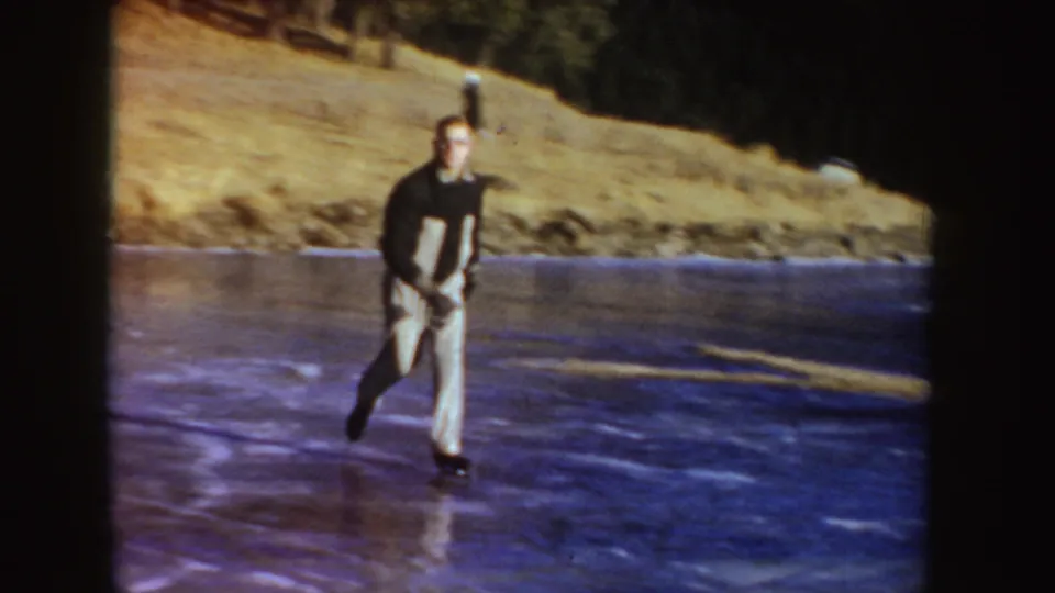 In 1955 Idaho home-movie footage, a man in a dark top and light pants skates across a frozen pond near a rural bank.