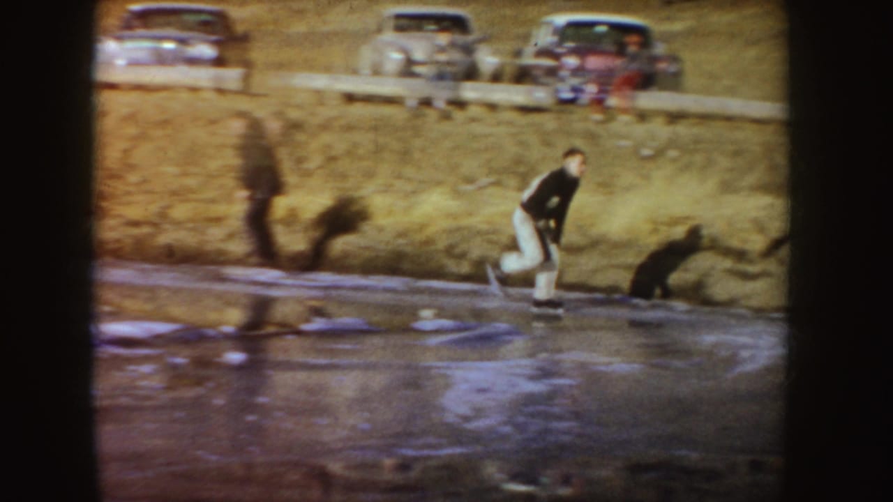 A man skates along the pond edge below parked mid-century cars on a grassy bank in Idaho home-movie footage.