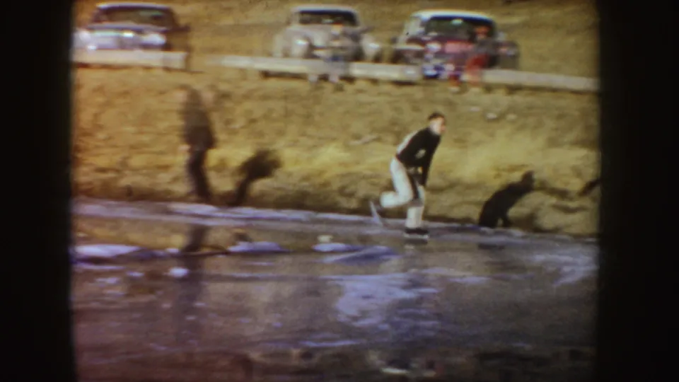 A man skates along the pond edge below parked mid-century cars on a grassy bank in Idaho home-movie footage.