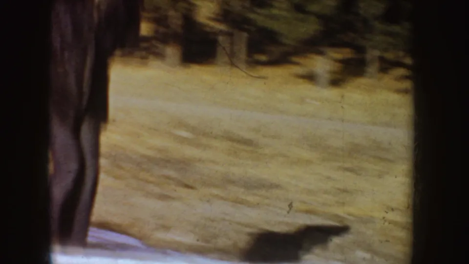 1955 Idaho home-movie frame showing the skater's legs beside the frozen pond surface and blurred tree line.