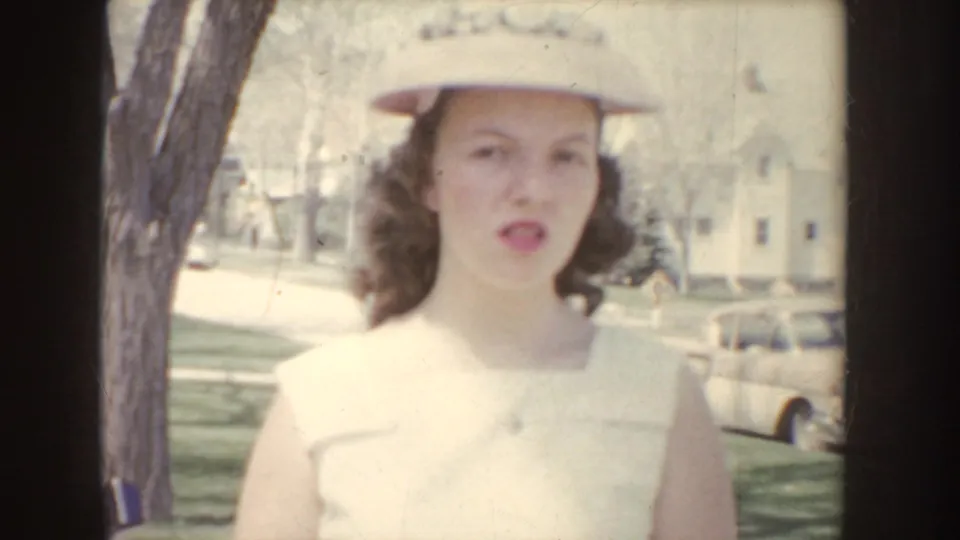 Young woman in a white dress and brimmed hat walks toward the camera on an Idaho residential sidewalk in 1955.