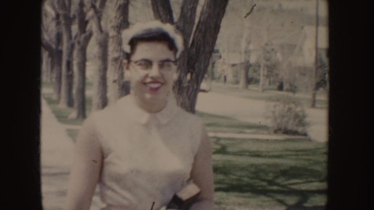 Smiling young woman in glasses and a white dress faces the camera beside trees and lawns in 1955 Idaho home-movie footage.