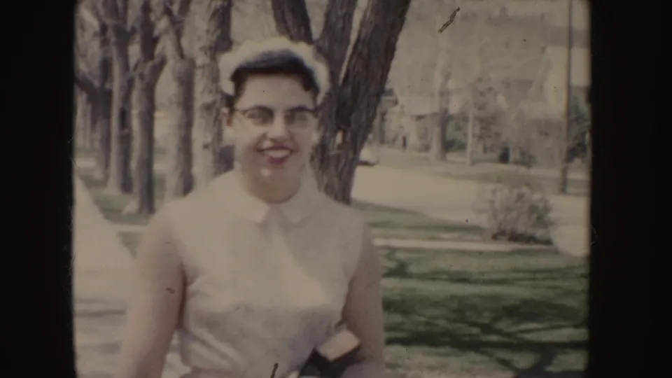Smiling young woman in glasses and a white dress faces the camera beside trees and lawns in 1955 Idaho home-movie footage.