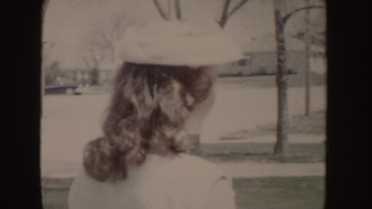 Rear view of a young woman in a white hat and dress walking away along a residential sidewalk in 1955 Idaho footage.