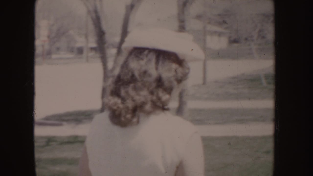 Soft 8mm frame of a woman in a white dress and hat from behind, with lawn, trees, and houses consistent with the Idaho 1955 metadata.
