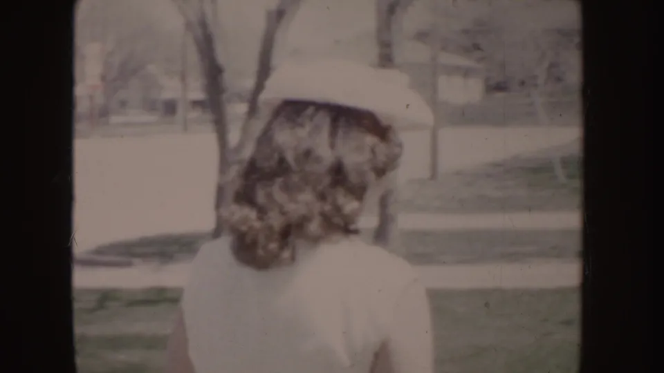 Soft 8mm frame of a woman in a white dress and hat from behind, with lawn, trees, and houses consistent with the Idaho 1955 metadata.