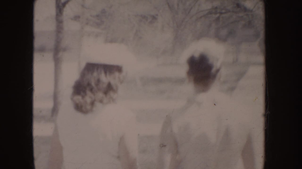 Faded 8mm frame showing two adults in light formal clothing moving through a residential outdoor setting in the 1955 Idaho clip.