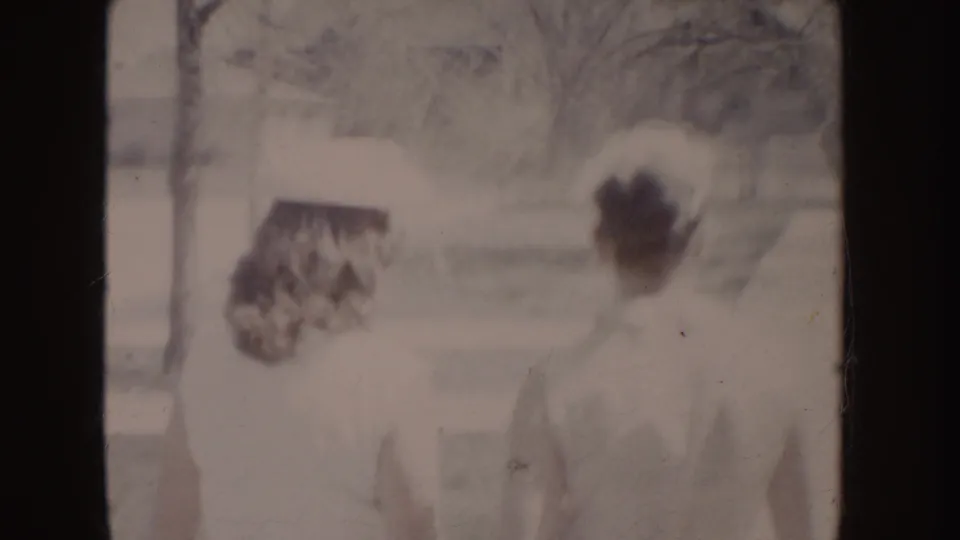 Faded 8mm frame showing two adults in light formal clothing moving through a residential outdoor setting in the 1955 Idaho clip.