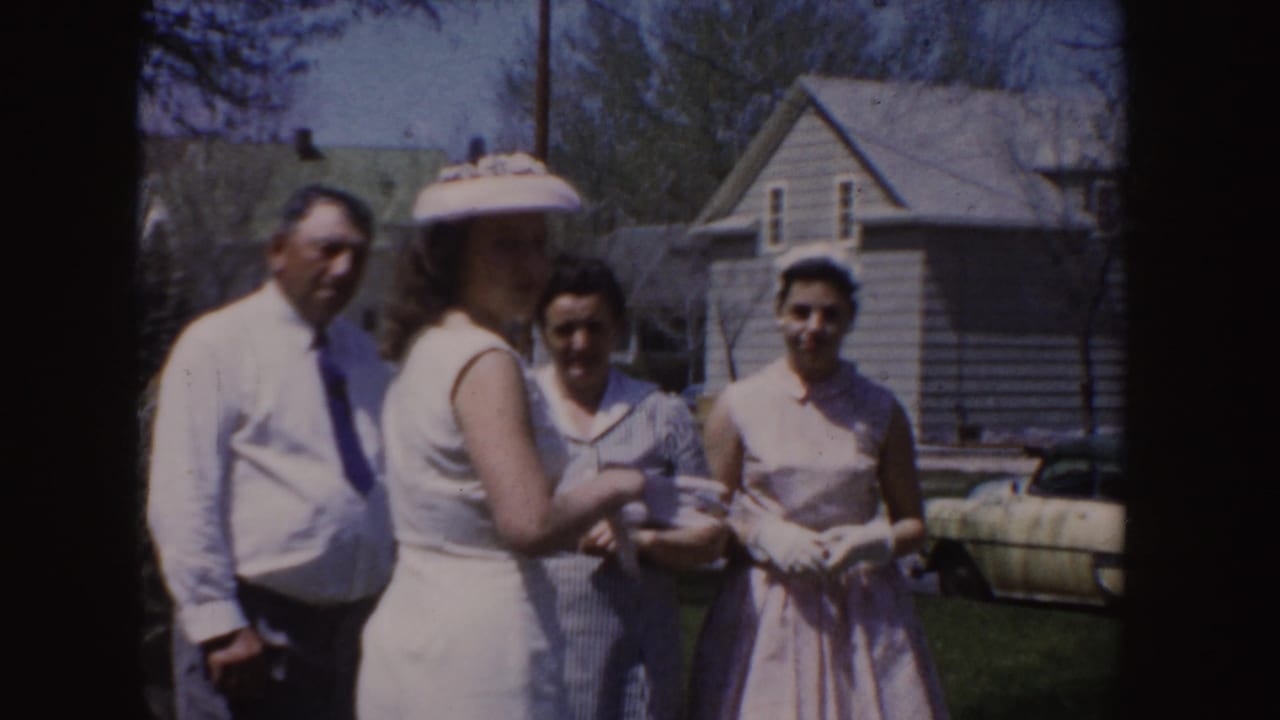 1955 Idaho home-movie frame with adults in dress clothes, including a man in a tie and women in light dresses, gathered near houses and a parked car.
