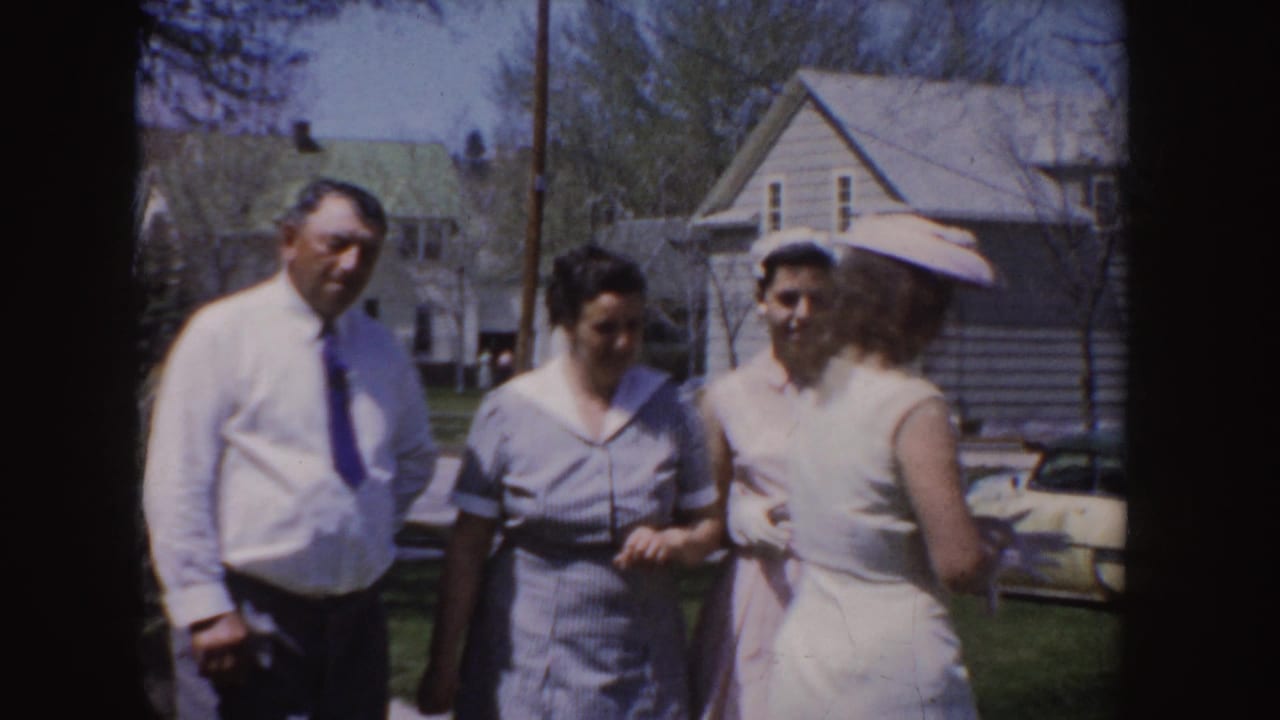 End frame of the 1955 Idaho 8mm clip showing adults in light dress clothing conversing in a suburban yard with houses and a parked car visible.