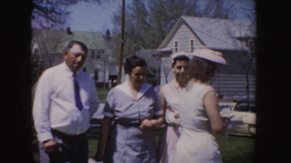 End frame of the 1955 Idaho 8mm clip showing adults in light dress clothing conversing in a suburban yard with houses and a parked car visible.