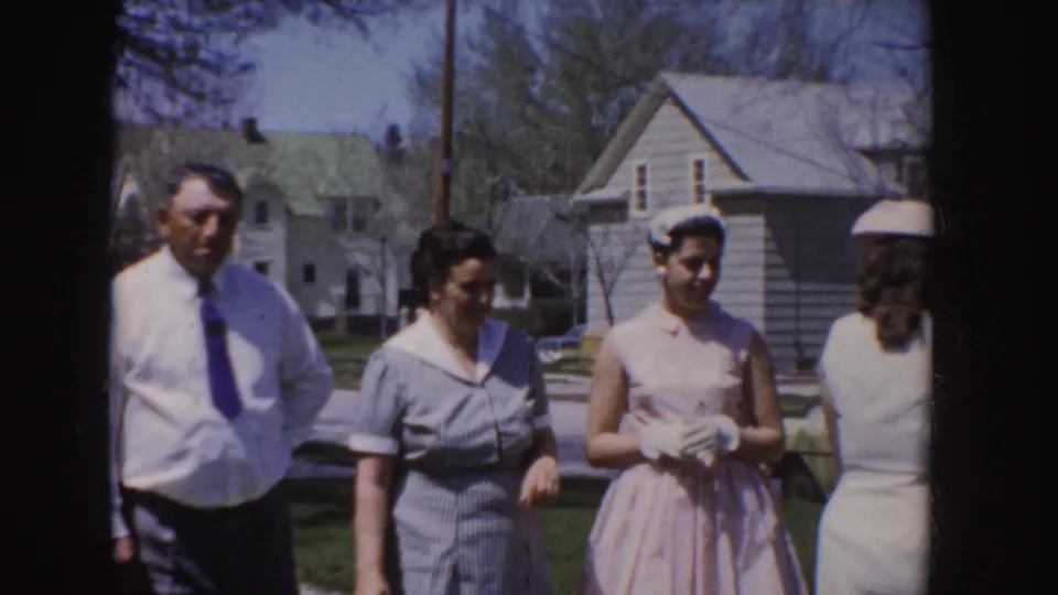 1955 Idaho home-movie frame showing a man in a white shirt and tie with women in dresses and hats outside residential houses.