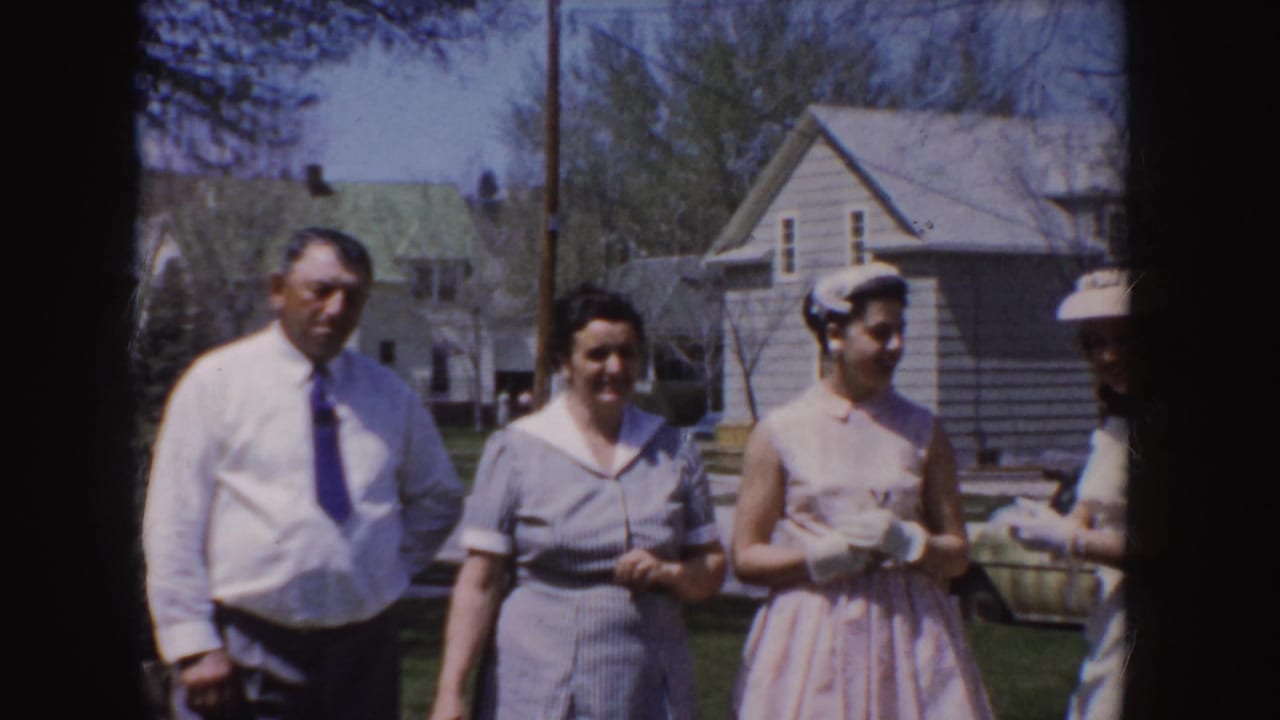 1955 Idaho outdoor frame with a man in a white shirt and women in mid-century dresses standing in a residential neighborhood.