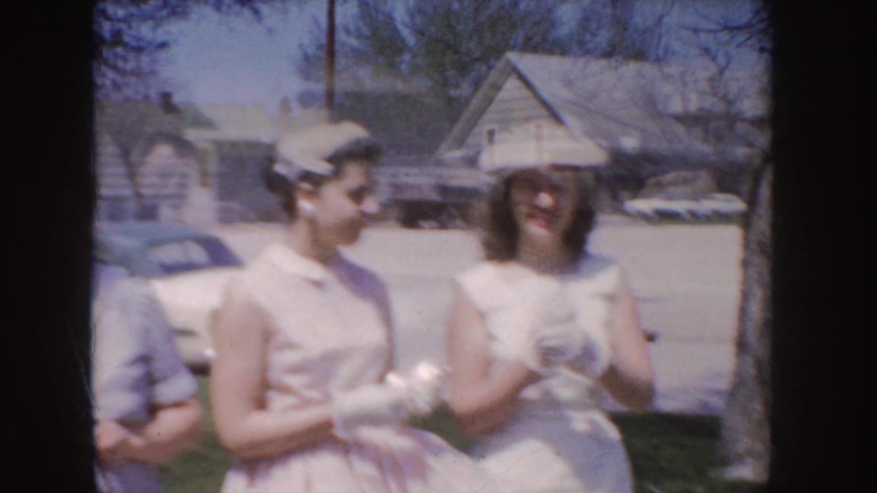 Soft 1955 Idaho home-movie frame of women in white and pink dresses with hats and gloves near a parked car and houses.