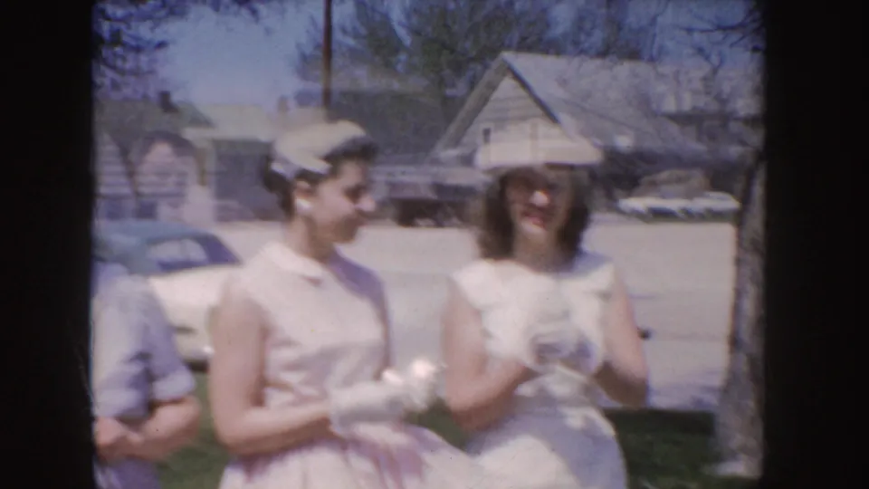 Soft 1955 Idaho home-movie frame of women in white and pink dresses with hats and gloves near a parked car and houses.