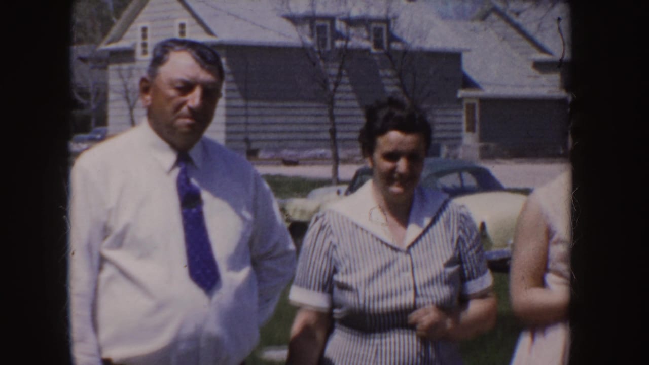 1955 Idaho frame showing a man in a white shirt and blue tie beside a woman in a striped dress, with a car and houses behind them.