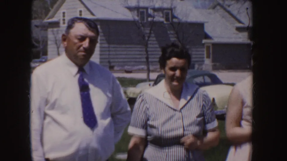1955 Idaho frame showing a man in a white shirt and blue tie beside a woman in a striped dress, with a car and houses behind them.