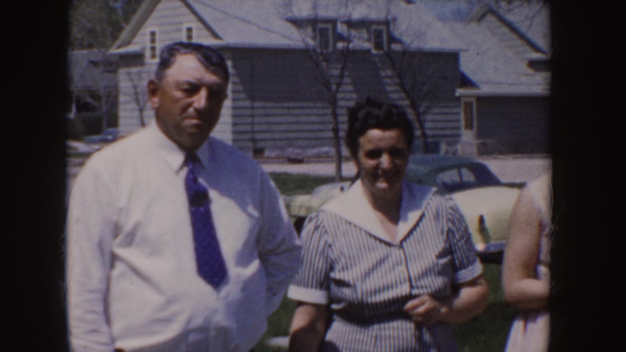 Man and woman stand outdoors near houses and a parked car, partly obscured by the film frame edge.