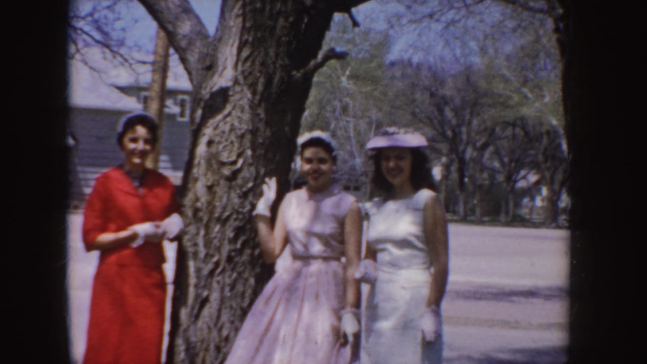 Three young women in 1950s formal dresses pose beside a large tree on an Idaho residential street.
