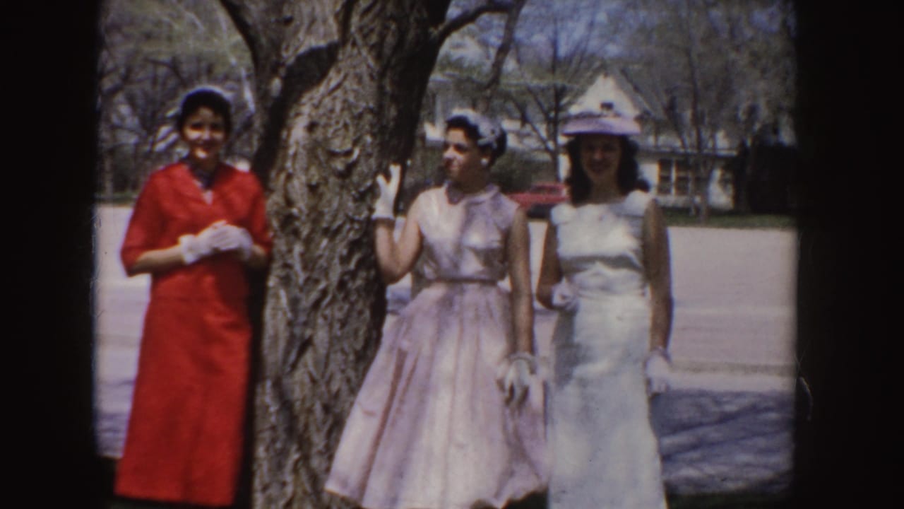 Three young women in red, pink, and white formal dresses pose together beside a tree in 1955 Idaho footage.