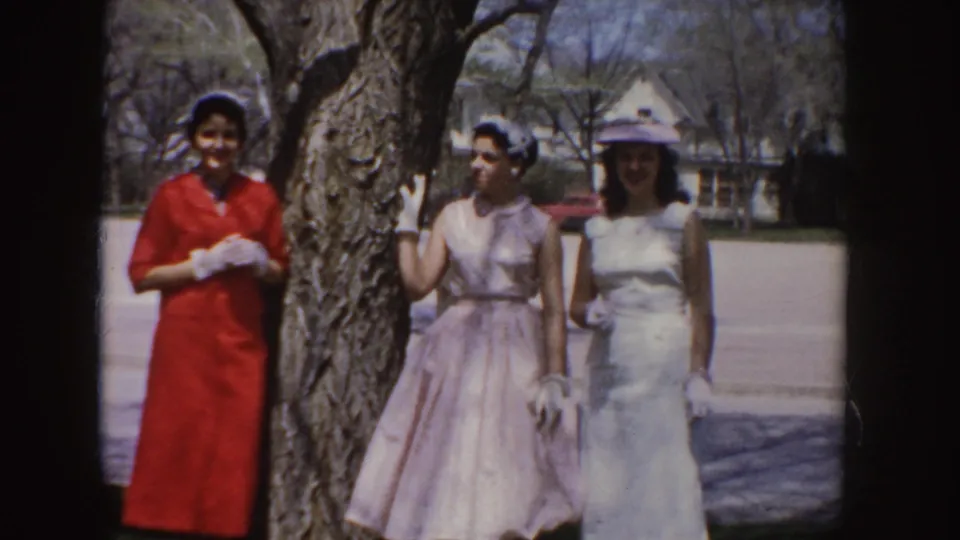 Three young women in red, pink, and white formal dresses pose together beside a tree in 1955 Idaho footage.
