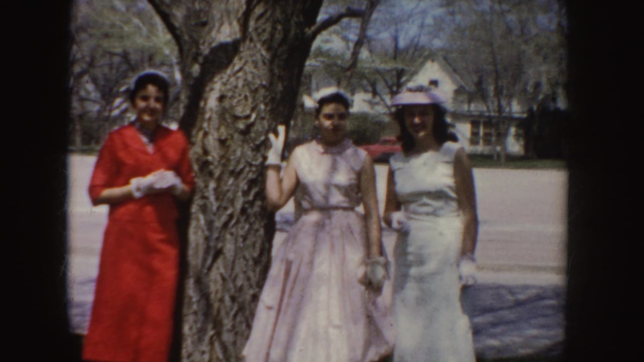 In a 1955 Idaho outdoor home-movie frame, three women in formal dresses smile near a tree with suburban houses behind them.