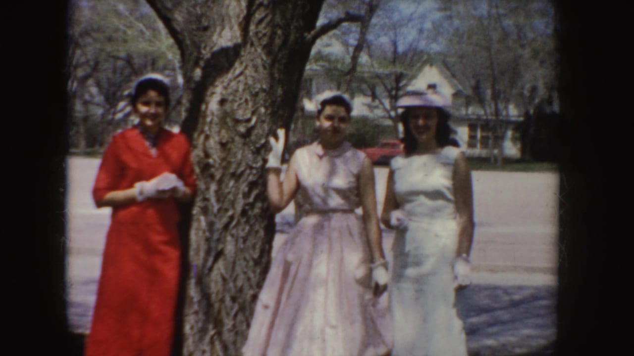 Three women in red, pink, and white dresses pose for the 8mm camera in Idaho, with gloves, a hat, and houses in the background.