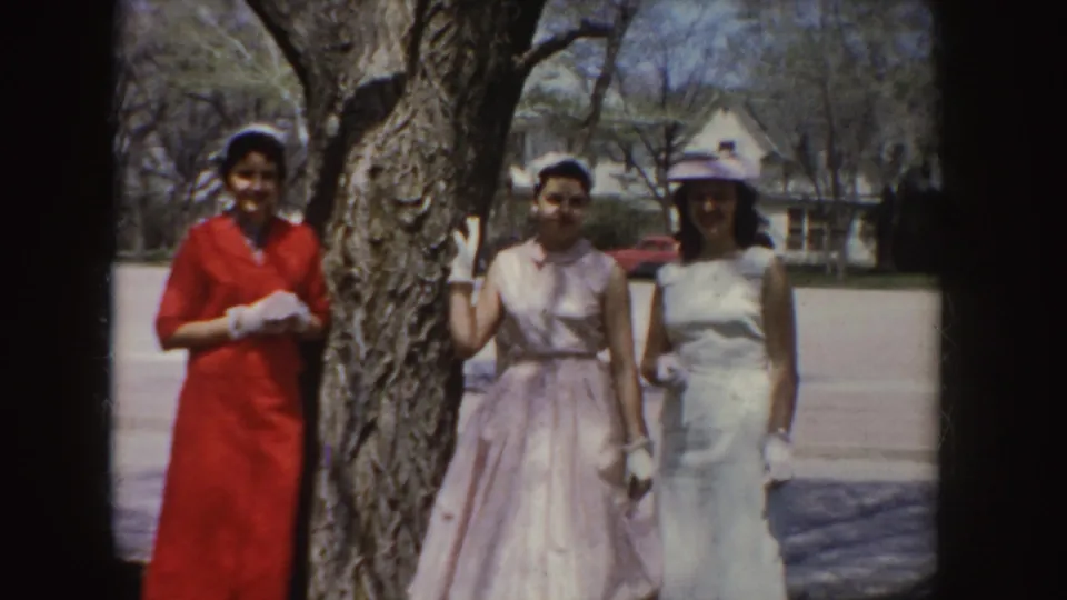 Three women in red, pink, and white dresses pose for the 8mm camera in Idaho, with gloves, a hat, and houses in the background.
