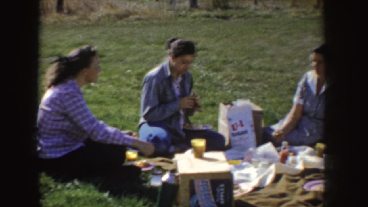 Three women sit together on grass with picnic items spread on a blanket.