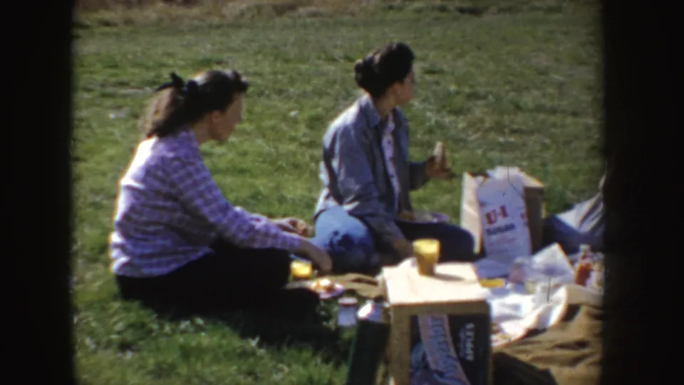 Home-movie frame of women seated at a picnic in Idaho, with a blanket, cups, food items, and a U-Haul bag visible.