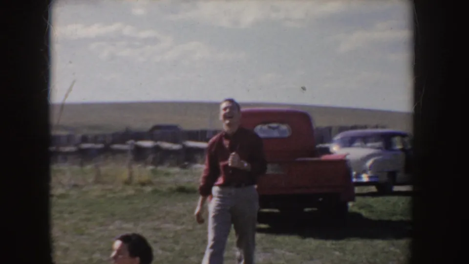 1955 Idaho frame showing a man in a red shirt near a red pickup and white car, with open land and sky behind him.