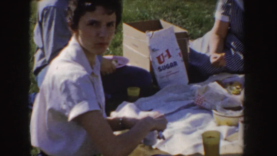 1955 Idaho home-movie frame showing a young woman at a park picnic with a U-I Sugar bag, food, cups, and other seated adults nearby.
