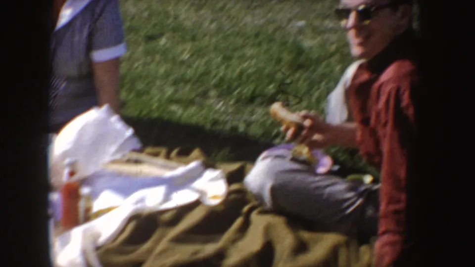 1955 Idaho home-movie frame of a woman in sunglasses seated on grass at a picnic, with blanket, food, and another seated adult nearby.