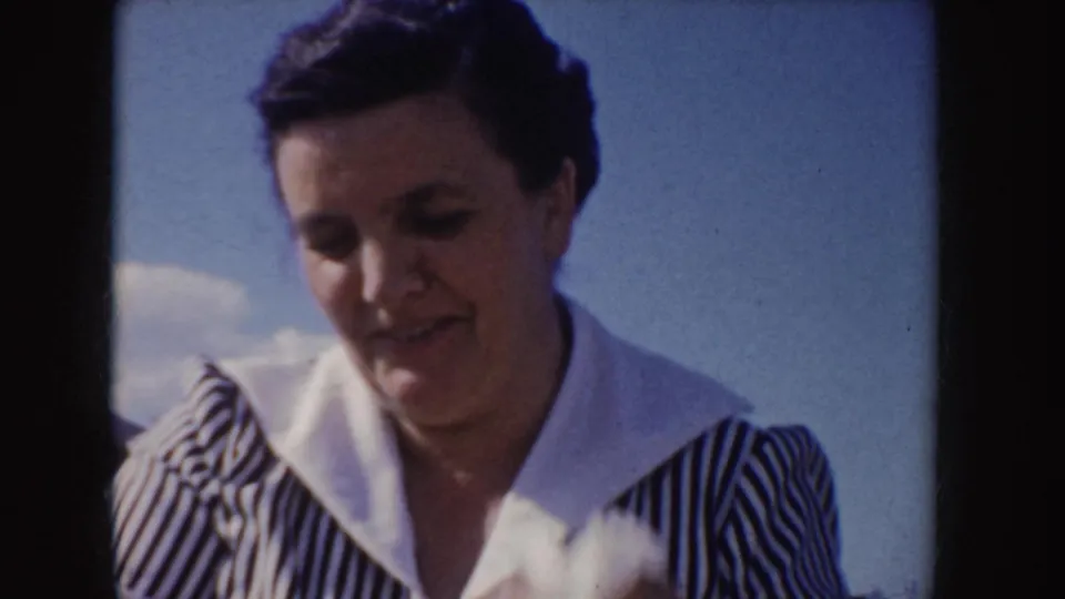 Close 1955 Idaho home-movie frame of a smiling woman in a blue and white striped dress with a broad white collar against the sky.