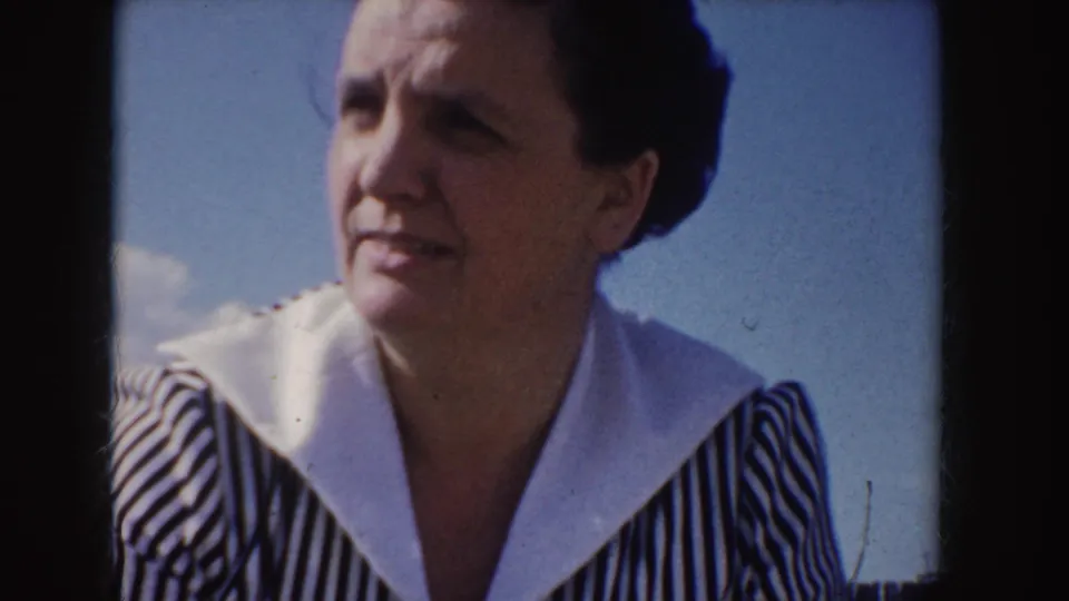 Adult woman in a striped dress and white collar turns her face left in 1955 Idaho home-movie footage.