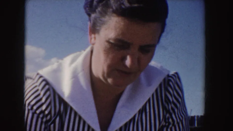 Close-up of an adult woman in 1955 Idaho footage looking down, framed by dark 8mm film borders and outdoor sky.