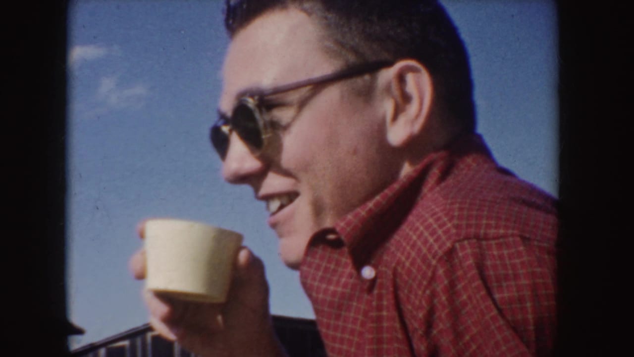 1955 Idaho frame of a man in dark-rimmed sunglasses and a red plaid shirt smiling with a light cup in his hand.
