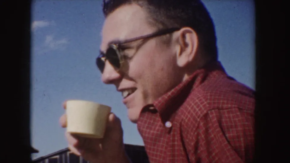 1955 Idaho frame of a man in dark-rimmed sunglasses and a red plaid shirt smiling with a light cup in his hand.