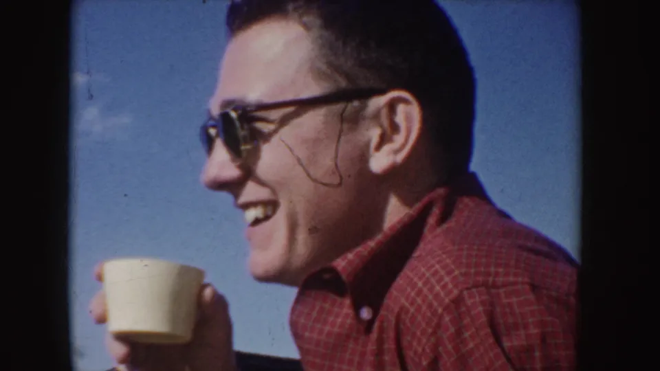 1955 Idaho home-movie frame showing the man in sunglasses laughing in profile with a cup near the edge of the frame.