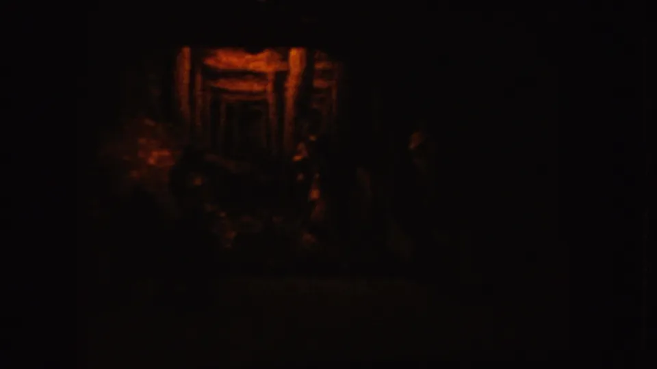 The low-light 8mm frame shows a warm orange-lit wooden form, possibly slats or a small structure, surrounded by black shadow.