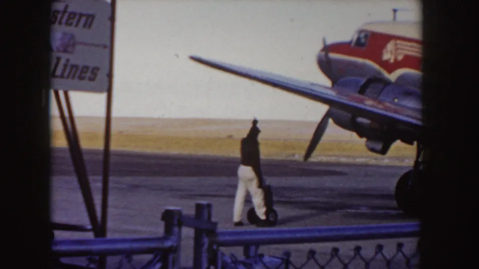 A ground crew figure walks near the wing and propeller area of the Western Airlines aircraft, with a fence and airline sign visible at the edge of the tarmac.