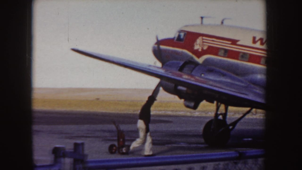 1955 Idaho 8mm frame showing the front section of a Western Airlines propeller plane on the tarmac, with a ground crew member and small cart near the wing.